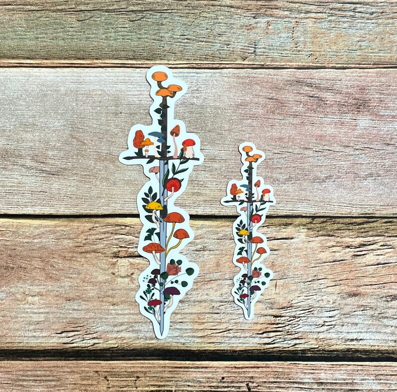 May include: Two stickers featuring a sword design with colorful mushrooms and foliage. The larger sticker is approximately 8 inches tall, while the smaller one is about 4 inches tall. The stickers have a white border and are set against a wooden background.