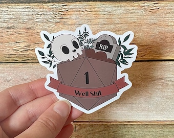May include: A white sticker with a brown dice with the number 1 on it. The dice is surrounded by green leaves and a white skull. The sticker has a red banner with the text "Well Shit" on it. The sticker also has a small tombstone with the text "RIP" on it.