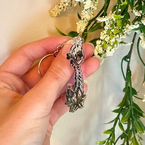May include: A silver key charm with a floral design. The key has a detailed design and is attached to a silver ring.