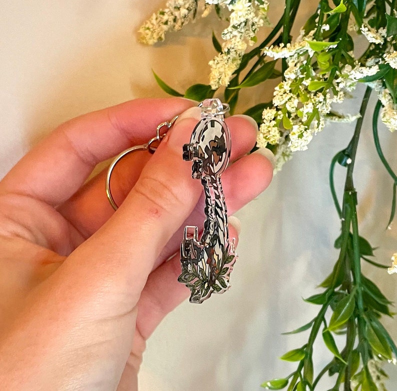 May include: A silver key charm with a floral design. The key has a detailed design and is attached to a silver ring.