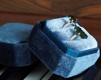 May include: Two rings with green gemstones set in silver are displayed in a blue velvet ring box. The box is open and the rings are visible. The box is sitting on a wooden surface.
