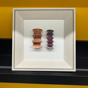 May include: Two small, framed sculptures, one in shades of copper and the other in shades of purple, are displayed against a white background.