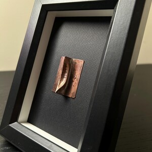 May include: A small, copper-coloured sculpture of a boat is framed in a black frame with a white mount. The sculpture is mounted on a black background.