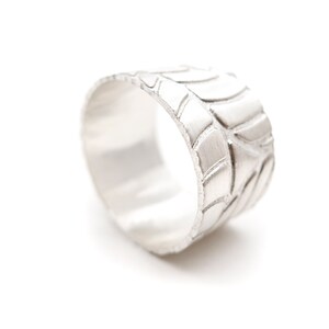 Puede incluir: Un anillo de plata con un patrón de banda de rodadura de neumático. El anillo es ancho y tiene un acabado liso y pulido.