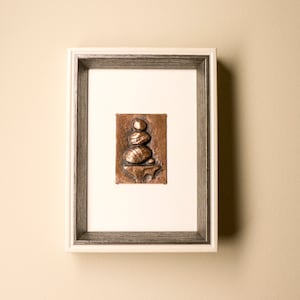 May include: A framed copper sculpture of a stack of stones on a base. The sculpture is a light brown colour and is set against a white background.