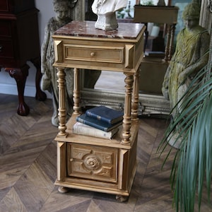 May include: A gold-coloured, antique-style side table with a marble top and a drawer. The table has ornate carved details and turned legs. Two shelves hold books. A white bust sculpture sits on top. The table is on a wooden floor.