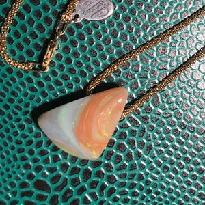 May include: A gold-toned necklace with a pendant featuring a triangular, multi-colored stone. The stone displays shades of white, orange, and light green. The necklace rests on a textured, teal-colored surface.