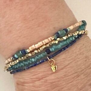 May include: A stack of four bracelets with blue, green, and gold beads. The bracelets are layered on a person's wrist and have a small gold charm hanging from the bottom.