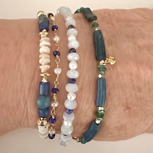 May include: A set of four bracelets featuring different colored beads. The bracelets are made with white, blue, and green beads. The bracelets are stacked on a person's wrist.