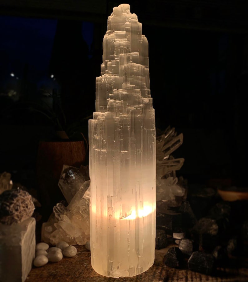 May include: A tall, white selenite crystal tower with a layered, textured appearance. The crystal is illuminated from within, creating a soft, warm glow. Other crystals and stones are visible in the background, creating a natural, spiritual aesthetic.