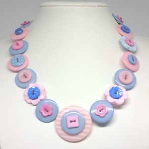 Pode incluir: Um colar de botões rosa pastel e azul com botões de vários tamanhos, incluindo alguns com desenhos florais.
