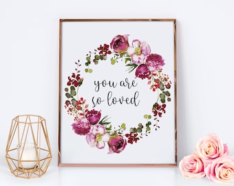 Könnte beinhalten: Ein gerahmter Blumendruck mit einem Aquarellkranz aus burgunderroten und rosa Blumen und grünen Blättern. Der Text "you are so loved" steht in der Mitte. Ein goldener geometrischer Kerzenhalter und rosa Rosen sind im Vordergrund.