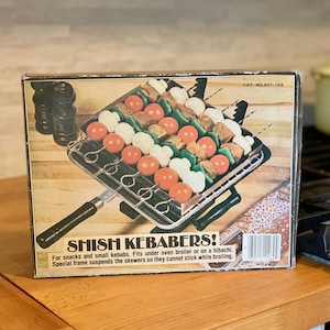 May include: A box for a shish kebab cooking device. The box is brown with a picture of the device on the front. The device is silver and has a black handle. The device is filled with vegetables and meat on skewers. The box says "Shish Kebabers!" and "For snacks and small kebabs. Fits under oven broiler or on a hibachi. Special frame suspends the skewers so they cannot stick while broiling."