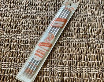 May include: A package of Susan Bates Silvalume knitting needles in a clear carrying case. The needles are silver and the packaging has orange and white text and a cartoon image. The needles are on a woven, light brown surface.
