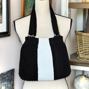 May include: A black and white crochet handbag with shoulder straps. The bag features a central white panel flanked by black sections. The straps are also black and attached to the bag with silver-colored rings. The bag is displayed on a mannequin.