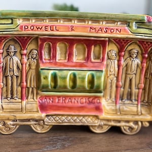 May include: A ceramic figurine of a San Francisco cable car with the words "Powell & Mason" and "San Francisco" on the front. The figurine is painted in shades of green, red, yellow, and brown. There are small figures of people on the car.