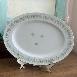 May include: An oval white porcelain serving platter featuring a delicate border pattern of small blue and pink flowers with green leaves. The center of the platter displays a small cluster of three matching floral sprigs on a smooth surface.