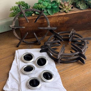 May include: An assortment of vintage stove parts, including black burner grates and silver and black control knobs, are arranged on a wooden surface. A white cloth is under the knobs. Decorative succulents are in the background.