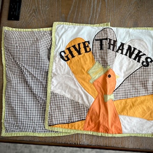 May include: A brown and white checkered throw pillow with a green border. The front of the pillow features a colorful applique of a turkey with the words "Give Thanks" in black.