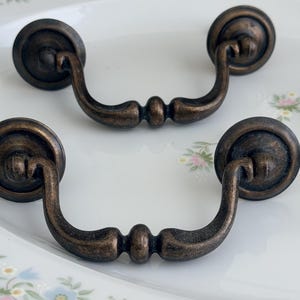 May include: A set of dark bronze-toned metal drawer pulls featuring a curved, swinging handle design attached to two circular mounting plates. The hardware has a weathered, textured finish with a central decorative detail on the handle.