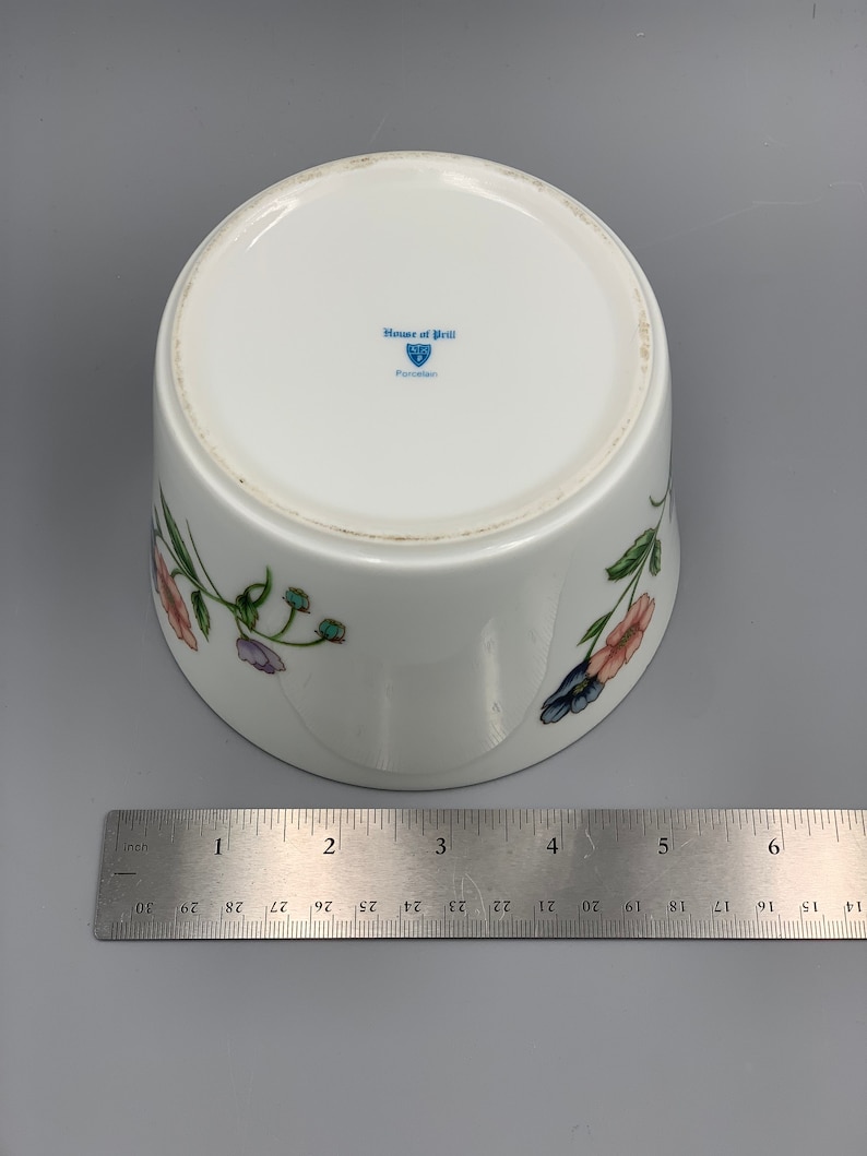 Puede incluir: Taz&oacute;n de porcelana blanca con un dise&ntilde;o floral con flores rosas, azules y verdes. El taz&oacute;n tiene un logotipo azul y blanco en la parte inferior que dice "House of Prill Porcelain".