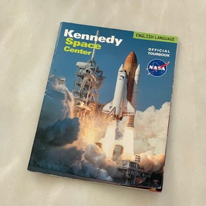 May include: A book titled "Kennedy Space Center" with a photo of a space shuttle launching. The book is an official tourbook in English and features the NASA logo.