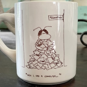 May include: White ceramic coffee mug with a cartoon illustration of a person overwhelmed by paperwork. The text on the mug reads "...wish I had a computer..." and "Accounting".