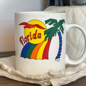May include: White ceramic mug featuring a colorful Florida design. The design includes a yellow sun, a rainbow, a palm tree, and the word "Florida" in red and yellow. The mug has a curved handle.