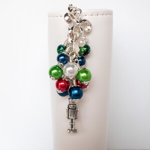 Peut inclure: Ornement de Noël argenté avec un nœud, des perles colorées et un charm miniature en forme de casse-noisette. L'ornement présente des perles rouges, vertes, bleues et blanches, créant un design de vacances festif. Le charm est détaillé.