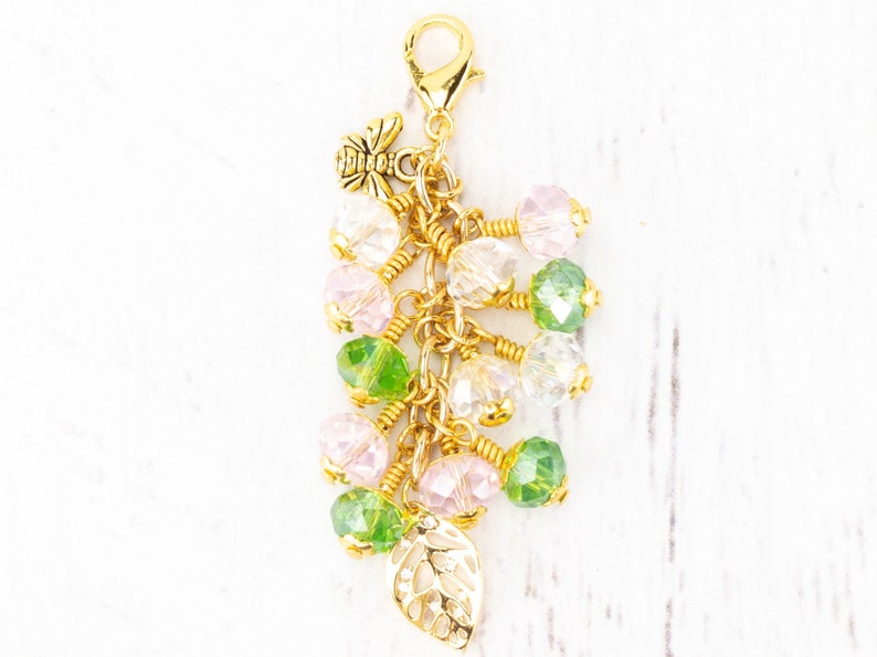Puede incluir: Un charm dorado con un cierre de mosquet&oacute;n, un peque&ntilde;o charm de abeja y un grupo de cuentas. Las cuentas son en tonos rosa, verde y transparente, con un charm de hoja dorada en la parte inferior. Para pulsera o collar.