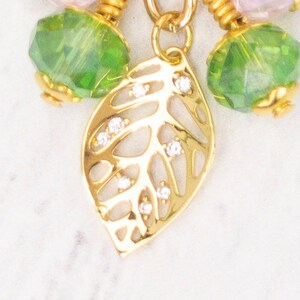 Puede incluir: Primer plano de un colgante de hoja dorada con peque&ntilde;as piedras preciosas transparentes. El colgante forma parte de un collar con cuentas facetadas verdes y rosas. La joya est&aacute; sobre un fondo blanco.