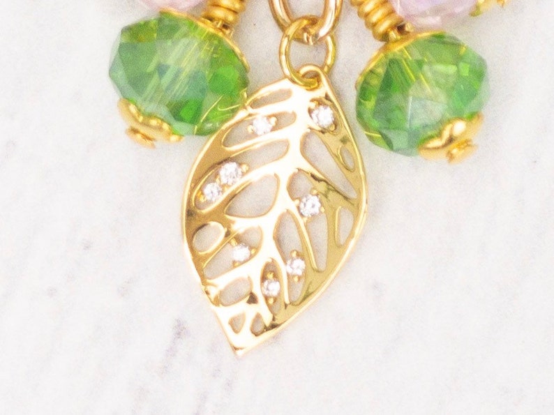 Puede incluir: Primer plano de un colgante de hoja dorada con peque&ntilde;as piedras preciosas transparentes. El colgante forma parte de un collar con cuentas facetadas verdes y rosas. La joya est&aacute; sobre un fondo blanco.