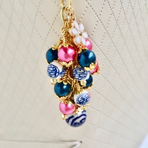 May include: A gold-toned charm with a cluster of beads in pink, navy blue, and white with blue patterns. A small white flower with gold accents is also attached. The charm is designed to be attached to a bag or keychain.