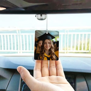 May include: Rectangular car air freshener featuring a smiling graduate in a black cap and yellow gown. The air freshener hangs from a silver chain, with a blurred background of a car interior and a scenic view.