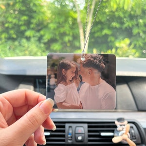 May include: Rectangular car air freshener with a printed photograph of a man and a girl. The photo is in color and shows the subjects smiling at each other. The air freshener hangs from a silver chain.