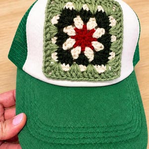 May include: A green and white trucker hat featuring a square crochet granny square patch on the front panel. The patch displays a floral-inspired pattern in shades of red, cream, and dark green, stitched onto the white foam front of the cap.
