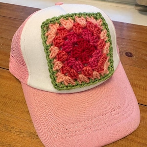 May include: A pink and white trucker hat featuring a square crochet granny square patch on the front panel. The patch is composed of concentric layers of yarn in shades of red, dark pink, light pink, and a sage green border.