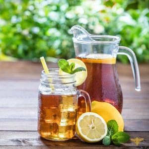 May include: A glass pitcher and mason jar filled with iced tea, garnished with lemon slices and mint leaves. The pitcher has a handle and a spout. The mason jar has a straw and a handle. Both containers are sitting on a wooden surface.
