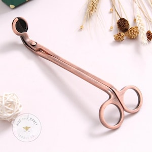 May include: A copper-toned candle wick trimmer with a rounded tip and a pair of scissors. The trimmer is designed to trim candle wicks to prevent soot and improve the burn.