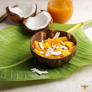May include: A coconut bowl filled with a tropical fruit salad. The salad includes pineapple, orange, and coconut. The bowl is sitting on a green banana leaf.