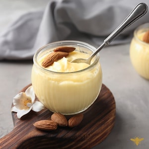 May include: A glass jar filled with a creamy yellow pudding topped with almonds. The jar is sitting on a wooden cutting board with additional almonds scattered around it. A silver spoon is resting on the rim of the jar.