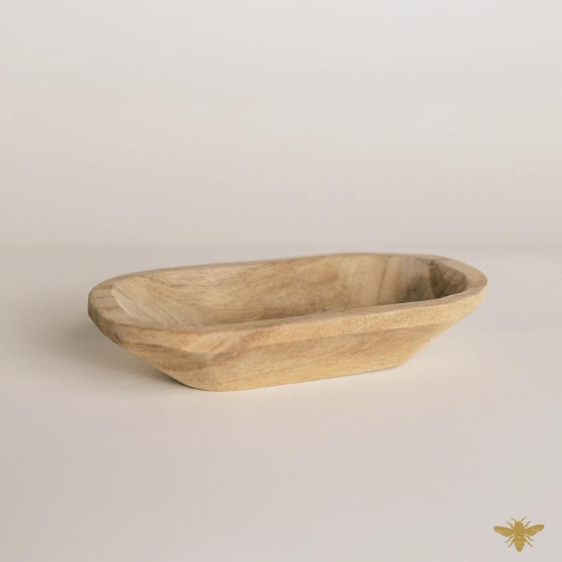 May include: A light-colored, wooden, oblong bowl with a shallow interior. The bowl has a smooth, natural finish and is likely hand-carved. The sides are gently sloped, and the overall shape is elongated. This decorative bowl is suitable for various uses.