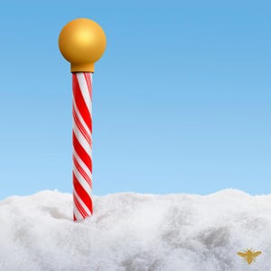 May include: A red and white candy cane with a gold ball on top is partially submerged in white snow.