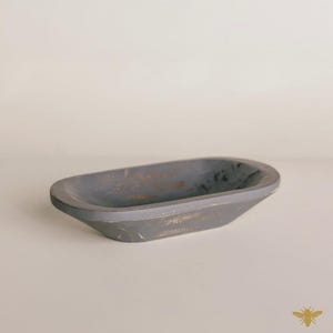 May include: A gray, oblong wooden bowl with a distressed finish. The bowl has a shallow, oval shape and is painted in a matte gray color with hints of gold. The bowl is likely used for decorative purposes or as a small serving dish.