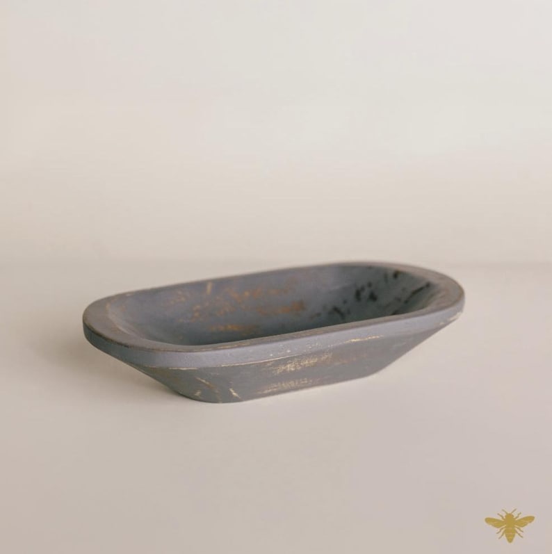 May include: A gray, oblong wooden bowl with a distressed finish. The bowl has a shallow, oval shape and is painted in a matte gray color with hints of gold. The bowl is likely used for decorative purposes or as a small serving dish.