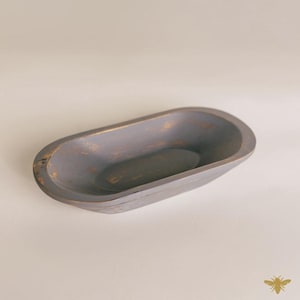 May include: A gray, oval-shaped wooden bowl with a distressed finish. The bowl has a shallow, elongated design and a slightly rough texture. The interior is a darker shade of gray, and the edges show hints of gold.