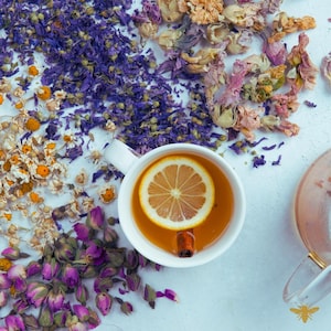 May include: A white teacup filled with tea, a lemon slice, and a cinnamon stick, surrounded by dried flowers in shades of purple, pink, yellow, and white. The tea is a warm amber color, suggesting a herbal infusion.