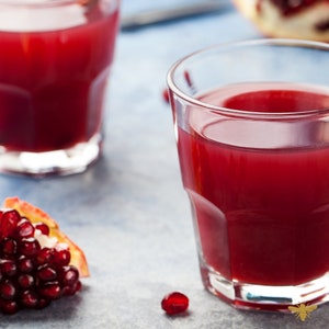 May include: Two glasses of red pomegranate juice on a blue background. A piece of pomegranate fruit is in the foreground.