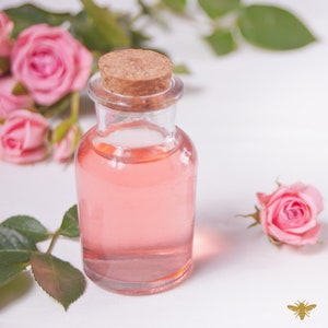 May include: A clear glass bottle with a cork stopper filled with a light pink liquid. The bottle is surrounded by pink roses and green leaves.