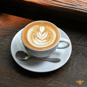 May include: A white ceramic mug with a latte art design on top of the coffee. The mug is on a white saucer with a silver spoon.